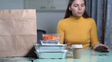 Paket servis menüsü. Bir kadın evdeki masada oturuyor ve teslimattan kalan yemekleri yiyor. Masaya birkaç adet tek kullanımlık plastik ve folyo tepsi konur. Seçici odaklanma. Paket yemek..