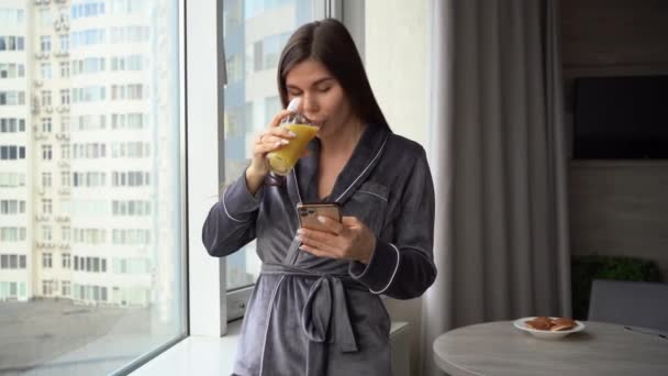 若い夢のような女性の肖像画は、彼女の手にオレンジジュースのガラスを保持し、彼女のスマートフォン上のソーシャルネットワーク上のニュースを読む朝に大きな窓の近くに立っている. — ストック動画