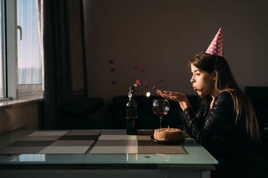 Evde izole edilmiş bir doğum günü, sofrada yalnız başına şapkalı bir kadın, yalnız bir tatilci, kek ve konfeti, şenlikli arka plan, içe dönük sosyal mesafe ve karantina konsepti.