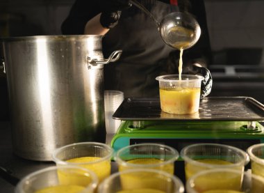 Restorana yemek teslimatı. Şef restoranda yemek hazırlar ve gerekli miktarı tartarak terazide tek kullanımlık tabaklar halinde hazırlar. Bir kepçenin yakın çekimi beslenme çantasına çorba döktü. Boşluğu kopyala.