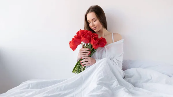 若い美しい白人女性が赤いチューリップの花束を手にした白いシャツを着てベッドの上に座っています 誕生日 バレンタインデー 記念日のためのギフトコンセプト コピースペース — ストック写真