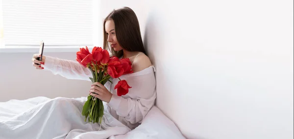 バレンタインデーのための寝室で朝に赤いチューリップの花束を持つセクシーな女の子の肖像画は 彼女のスマートフォンで自撮りを行います ベッドの上で花を持つ女性女性の日 コピースペース — ストック写真