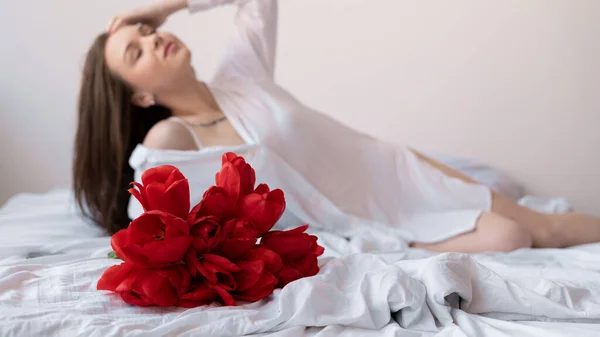 花とベッドの上のベッドルームでセクシーな女の子 白い背景に赤いチューリップを持つ女性は 朝の目覚めを楽しんでいます ホームコンセプトで幸せな週末 テキストの場所 — ストック写真