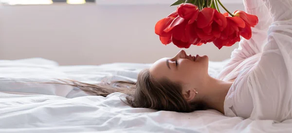 a young beautiful Caucasian woman lies on a bed in a white shirt holds red tulips in her hands and inhales their aroma. gift concept for birthday, valentine's day, anniversary. copy space.
