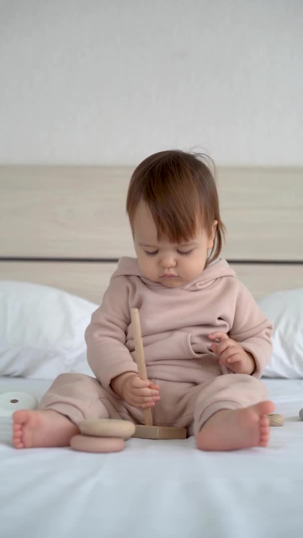 Cute baby girl gra z kolorowe tęczowa piramida zabawka siedzi na łóżku w białej, słonecznej sypialni. Zabawki dla małych dzieci. Dziecko z zabawką edukacyjną. pionowe wideo. Rozwój wczesny. — Wideo stockowe