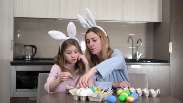 Pasen. familie moeder en dochter met haas oren zijn de voorbereiding voor de vakantie, verf eieren in een gezellige keuken thuis zitten aan de tafel. — Stockvideo