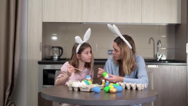 Semana Santa. familia mamá y su hija con orejas de liebre se están preparando para las vacaciones, pintar los huevos en una acogedora cocina en casa sentado a la mesa. comunicarse bien y pasar tiempo juntos. — Vídeo de stock