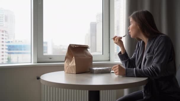 Una donna si siede a casa a un tavolo in pigiama e mangia cibo biologico dalla consegna. Il sacchetto di carta è sul tavolo. Scatola usa e getta con pellicola. — Video Stock