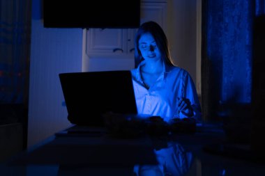 İnternette uzaktan çalışırken gece geç saatlerde evde yemek yemek. Teslimattan yemek yiyen ve dizüstü bilgisayarı üzerinde çalışan güzel bir kızın portresi. Boşluğu kopyala.