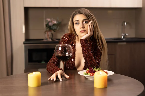 赤のドレスを着た美しい女性がワインを片手にテーブルに座り 悲しい表情でカメラを覗き込む 近くのテーブルにはろうそくがあります このコンセプトはバレンタインデーや記念日に孤独です スペースのコピー — ストック写真