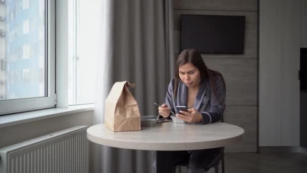 La chica se sienta en casa y come comida de la entrega en la mesa en una lonchera de papel aluminio desechable. Desplazamiento a través de las noticias de las redes sociales utilizando un teléfono inteligente. La emoción de la sorpresa. — Vídeo de stock