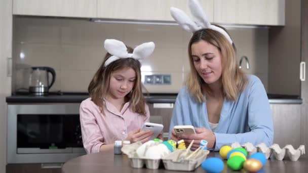 Easter family. mom and daughter with hare ears are preparing for the holiday, painting eggs in the cozy kitchen at home, sitting at the table and reading the news on their smartphones. — Stockvideo