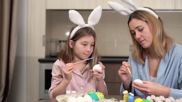 Pasen. familie moeder en dochter met haas oren bereiden zich voor op de vakantie, schilderen eieren in een gezellige keuken thuis zitten aan de tafel. communiceren en samen tijd doorbrengen. — Stockvideo