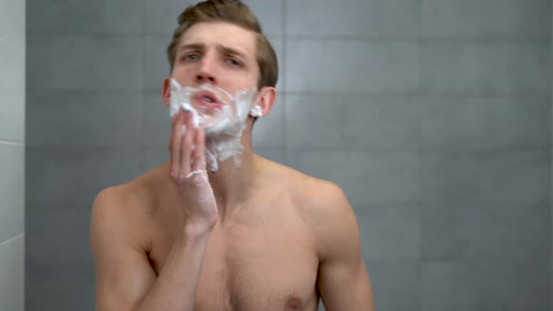 Positive attractive young guy applies helium foam in front of the mirror. A handsome man enjoys a morning bath, takes care of his appearance, healthy skin. Hygiene, skin care. — Wideo stockowe