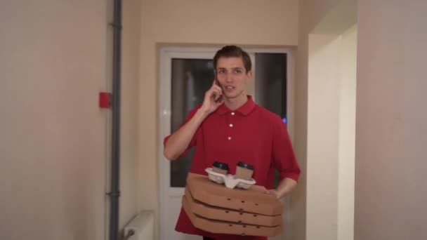 A pizza delivery man walks indoors along the corridor looking for an address and makes a call to the customer using his smartphone. Courier with food on the floor. — Stok video