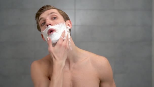 Красивий незахищений молодий чоловік наносить зволожуючу піну для гоління на обличчя і дивиться на камеру. Портрет щасливого чоловіка, що виконує щоденну особисту допомогу вдома у ванній — стокове відео