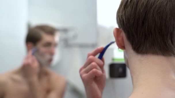 Close-up of the back of the head of a man looking in the bathroom mirror, shaving his face after a shower, a thousand-year-old man takes care of his stubble or beard, does a morning cosmetic procedure — Wideo stockowe