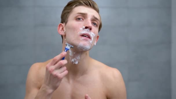 Joven hombre caucásico con espuma en su cara afeitándose con una navaja de afeitar, hombre milenario cuidando de rastrojos o barba, haciendo tratamiento facial matutino en baño, s — Vídeo de stock