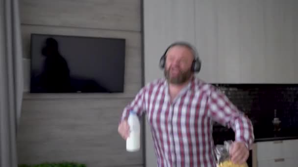 A bearded man in wireless headphones walks through the kitchen with a bottle of milk and dances. Bachelors breakfast milk and cornflakes. — Video