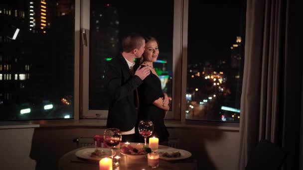 Una pareja enamorada, un hombre y una mujer se paran en la ventana por la noche y se abrazan en el fondo de la ciudad nocturna. — Vídeos de Stock