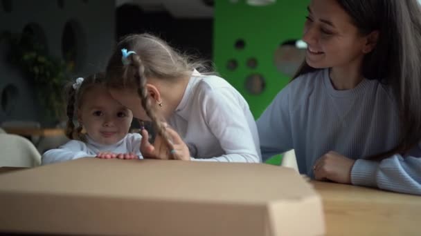 A woman with two children girls sits in a cafe sit at a table near a pizza box. Mom and two daughters eat together. — Vídeo de Stock