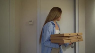 A woman has received an order, enters the room and holds several boxes of pizza in a food delivery box.