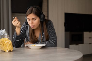 Happy woman holding bowl and spoon eating cereal while sitting at home. listen to music in wireless headphones during breakfast. copy space.