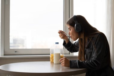 A young woman is sitting at a table near the window during breakfast. listens to music on headphones and eats corn flakes with milk. copy space.