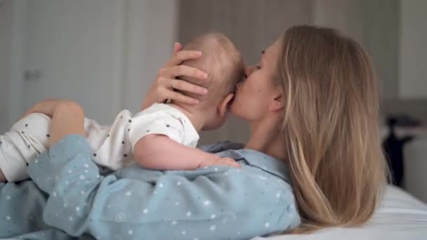 Όμορφη νεαρή λευκή γυναίκα ξαπλώνει στην πλάτη της και κρατά το μωρό της στο στομάχι της. Η μαμά αγκαλιάζει και φιλάει τον μικρό της γιο. Έννοια ημέρας μητέρων. — Αρχείο Βίντεο