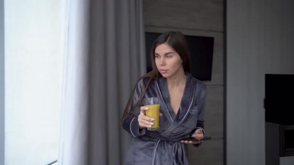 Young woman looking into smartphone while drinking orange juice in the kitchen at home. Woman with healthy food indoors. Morning routine. Young woman enjoying fresh orange juice, reading the news. — Stockvideo