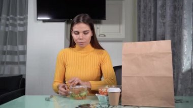 Masada tek başına oturan beyaz kadın içinde diyet salatası olan plastik bir konteynırı açar ve akşam yemeği yemeye başlar. Eve servis gıda konsepti.