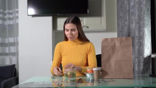 Conceito de comida entrega em casa. Uma jovem senta-se à mesa à noite, abre uma bandeja de plástico com comida e começa a comer uma salada de legumes.. — Vídeo de Stock