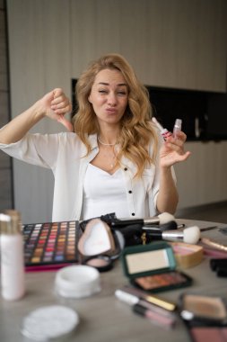Genç ve çekici beyaz ırktan modelin tripodla akıllı telefondan videoya alınıp makyaj malzemeleriyle masada ruj ve baş parmağını göstermesi. Kötü eleştiri ve mutsuz müşteri. portre