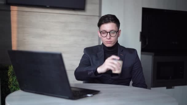 Homem de negócios trabalhando em casa com um laptop. senta-se em uma jaqueta e copos na mesa bebendo café de uma xícara descartável. O conceito de um trabalhador remoto trabalhando em um escritório em casa. — Vídeo de Stock