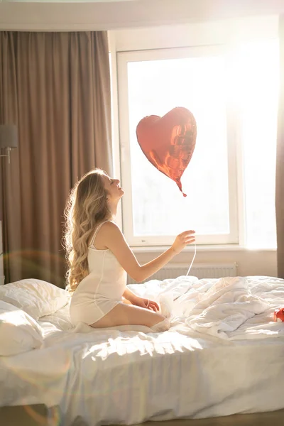 太陽の下で午前中にベッドの上に自宅で美しい若い女の子 赤いハート型の風船でバレンタインデーのお祝いをお楽しみください コピースペース — ストック写真