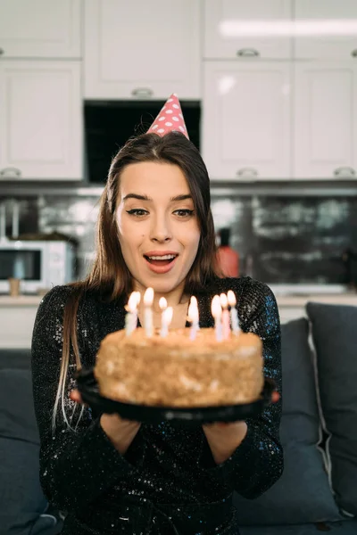 年轻女人在生日蛋糕上吹灭蜡烛 一个戴着派对礼帽的女孩吹灭蜡烛 在家里庆祝 快乐的生日理念 复制空间 — 图库照片