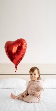 Sevgililer günü ya da doğum günü konsepti tatlı kız bebek ve evde yatakta oturan uçan kırmızı hava balonu. Boşluğu kopyala.