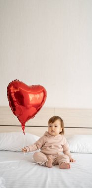 Küçük beyaz kız yatakta oturuyor ve elinde kalp şeklinde kırmızı bir balon tutuyor. Pankart. Boşluğu kopyala.