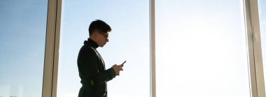 Erkek işadamı cep telefonları için cep telefonu kullanıyor panoramik pencerenin yanındaki ofiste duruyor. Dijital teknoloji uygulamaları ve kurumsal iş geliştirme için çözümler. Boşluğu kopyala.