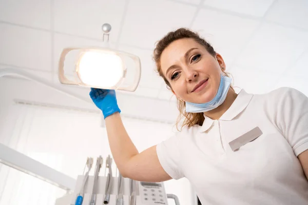 young female dentist smiling, wearing a medical mask, adjusting a dental lamp. A professional dentist is preparing for a dental examination, looking at the camera. POV of the patient. Medicine concept