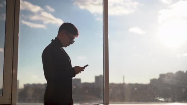 Бизнесмен в современном офисе ведет переговоры по мобильному телефону, обзванивает партнеров, владельца бизнеса перед большим окном небоскреба — стоковое видео