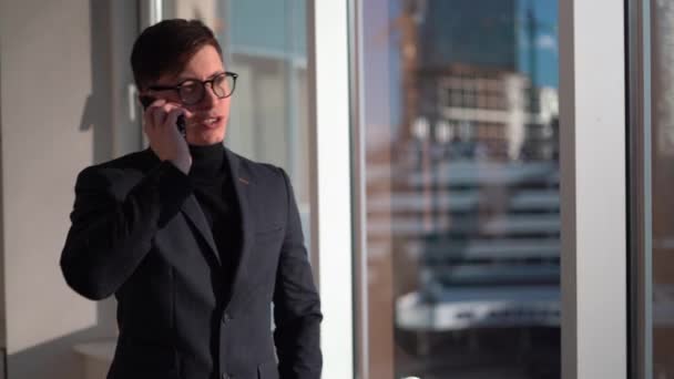Hombre de negocios caucásico en gafas hablando por teléfono mientras está de pie en la ventana, negociaciones de negocios en la oficina, llamada a socio de negocios — Vídeos de Stock