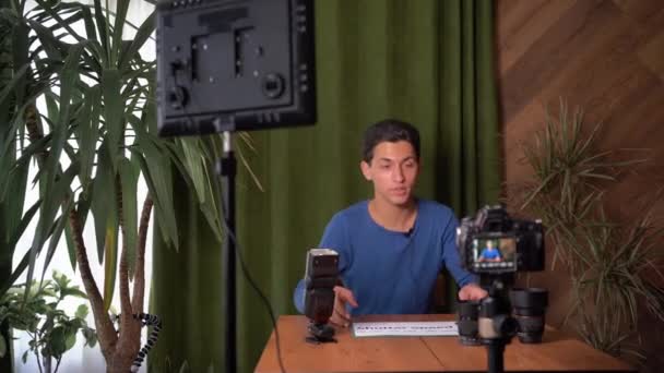 Aula de vídeo sobre como escolher o equipamento fotográfico e como trabalhar com ele. Um jovem muçulmano atraente está filmando a si mesmo na câmera. segura em suas mãos um sinal com a velocidade do obturador de texto. — Vídeo de Stock