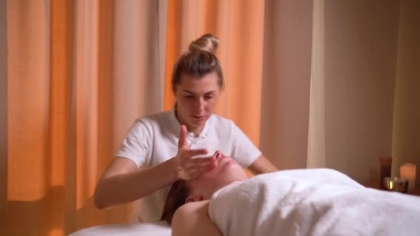 Esteticista masajista masajea la piel de una cara de mujer en una clínica de cosmetología. El esteticista comienza el masaje facial. Tratamientos de spa — Vídeo de stock