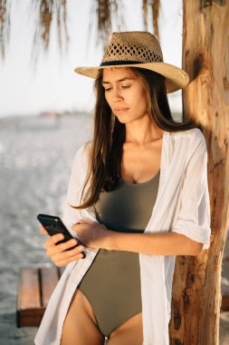 Denizde tatil, kumsalda hafta sonu, şapkalı bronzlaşmış güzel bir kız akıllı telefon ekranına bakıyor, saman şemsiyesinin altında sahilde duruyor. yakın plan portre