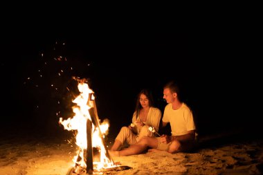 Deniz sahilinde genç bir çift ateşin yanında oturur ve çubuğun üzerinde marşmelov kızartırlar. Ateşin yanında romantik bir randevu. Marshmallow barbeküsü. Turizm ve turizm konsepti. Boşluğu kopyala.