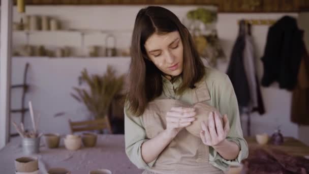 Mooie ambachtelijke vrouw die een kleikom maakt in een aardewerk werkplaats. Ze kijkt naar het product en glimlacht. Het concept van favoriete hobby en creatieve ontwikkeling — Stockvideo