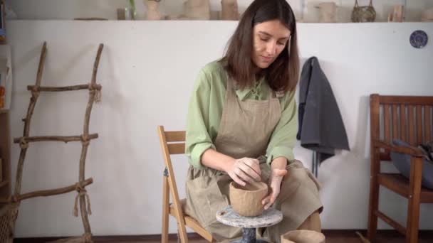 漂亮的年轻女性陶工塑造一个陶瓷杯,光滑,塑形和雕塑.那位女士坐在长椅上，手里拿着一个陶瓷轮，还做了一个陶罐. — 图库视频影像