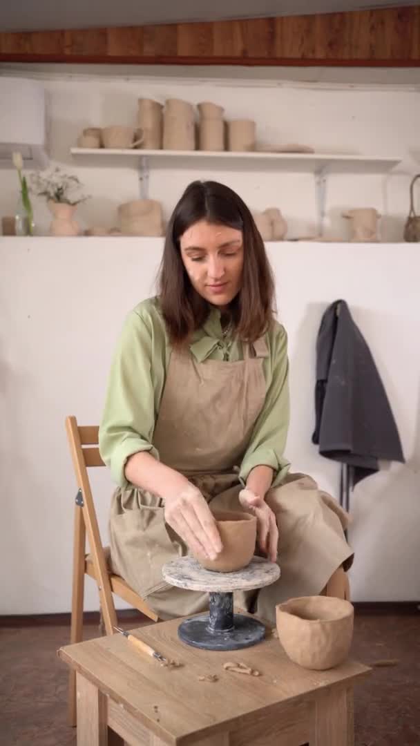 Piękna młoda garncarka kształtująca filiżankę ceramiki, wygładzająca, kształtująca i rzeźbiąca. Pani siedzi na ławce z kołem garncarskim i robi gliniany garnek. pionowe wideo. — Wideo stockowe