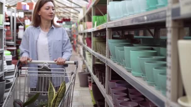 Kvinna väljer keramiska krukor för inomhusväxter i en trädgård centrum. En kvinna köper en kruka för att plantera växter hemma i ett lager. levereras med stormarknad kundvagn — Stockvideo